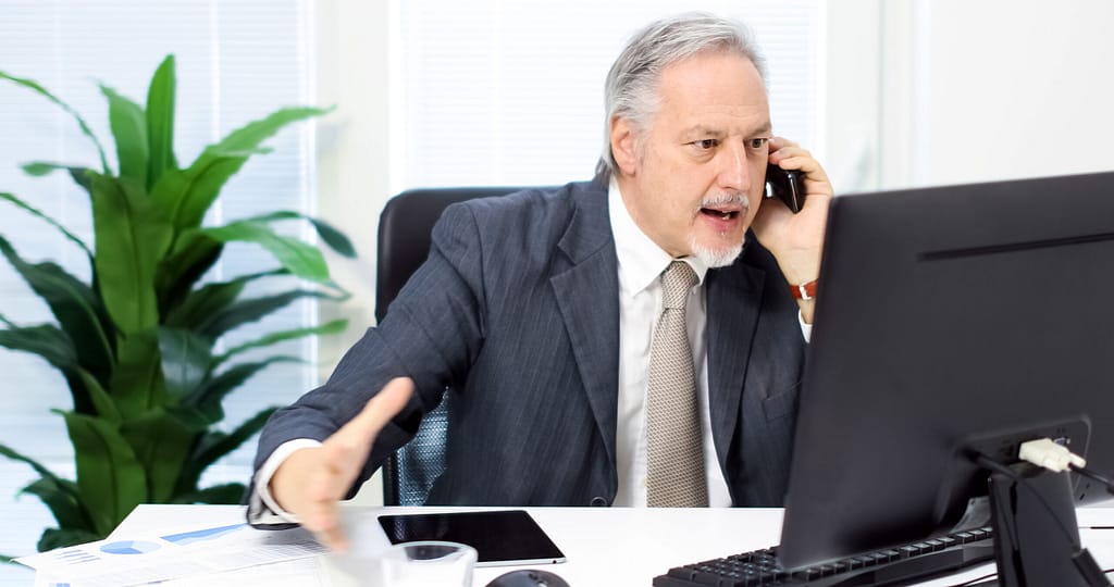 Angry man sitting at computer and talking on the phone.